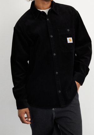Personne portant une chemise en velours côtelé noire à boutons avec une poche poitrine et un logo Carhartt, associée à un pantalon foncé, sur un fond uni.