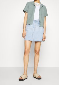 Chemise vert clair à manches courtes, t-shirt blanc, jupe en denim bleu clair avec ourlet effiloché, sandales beiges à semelles noires.