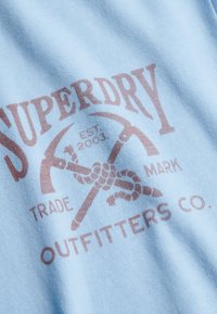 T-shirt en coton bleu clair avec un logo graphique marron montrant des pioches croisées et le texte « Superdry Outfitters Co. ».