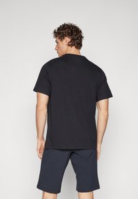 Tricou negru din bumbac, cu mâneci scurte, gât rotund și croială lejeră, asortat cu șorturi bleumarin, observat din spate.