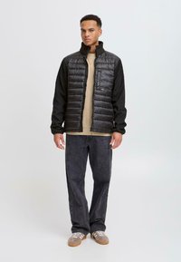 Svart dunvest med horisontale sømmer og høy krage, kombinert med en beige topp og mørkegrå vide jeans. Uformelt fottøy fullfører antrekket.