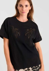 Camiseta de algodón negra con mangas cortas y enrolladas. Presenta dos diseños de palmeras en detalle recortado, destacando un ajuste casual y relajado.