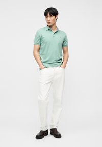 Νέος άνδρας όρθιος με τα χέρια στις τσέπες, φοράει μπλουζάκι πόλο σε μέντα πράσινο χρώμα, λευκό παντελόνι και σκούρα καφέ μπότες σε απλό φόντο.