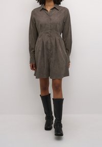 Robe-chemise marron à boutons avec manches longues et taille plissée, associée à des bottes noires montantes jusqu'au genou. Texture de tissu lisse, col classique.