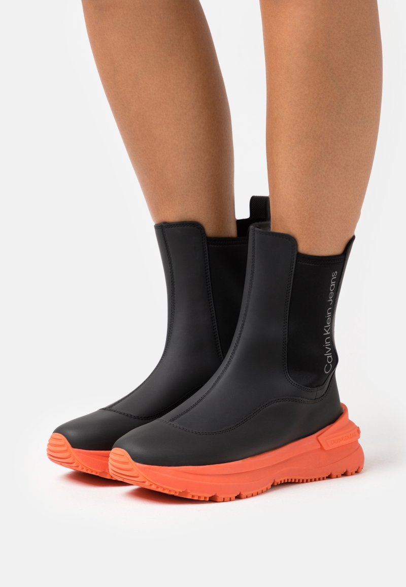 Czarne botki za kostkę o gładkim, matowym wykończeniu, z pomarańczową gumową podeszwą i małym detalem logo po boku.