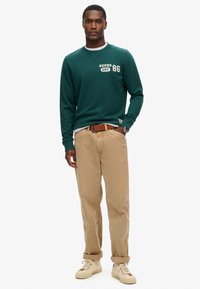Felpa verde con colletto bianco, testo stampato e logo; pantaloni beige; scarpe beige; modello in piedi su uno sfondo bianco.