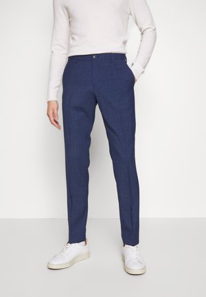 Pantalons bleus ajustés avec un motif à carreaux subtil, fabriqués dans un tissu léger, associés à un pull beige et des baskets blanches.