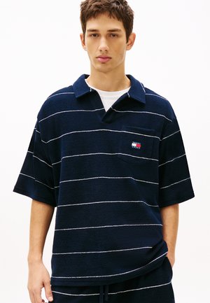 Młody mężczyzna w granatowej, krótkorękawowej polo w paski z logo Tommy Jeans na kieszeni na piersi.