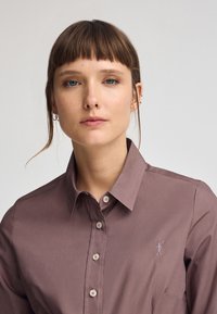 Camicia a manica lunga con colletto in mauve tenue e texture liscia. Presenta bottoni chiari e un piccolo design ricamato sul petto.