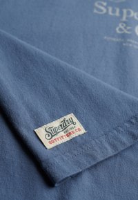 Blå bomuld T-shirt med hvidt logo og en vævet etiket med "Superdry" og "Outfitters Co." i røde og blå accenter.