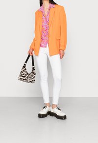 Oranžový blazer, růžová květinová košile s knoflíky, bílé úzké džíny, taška s černobílým zebrovaným vzorem a bílé boty s tlustou černou podrážkou.