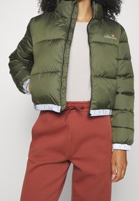 Giacca imbottita verde oliva con colletto alto, polsini elasticizzati e rifiniture in azzurro chiaro a contrasto, con logo sul petto.