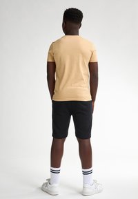 T-shirt beige clair à manches courtes, shorts noirs, baskets blanches et chaussettes blanches rayées, vues de l'arrière sur un fond uni.