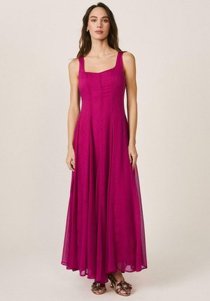 Mujer con cabello largo y castaño, vestida con un vestido fucsia sin mangas y de largo hasta el suelo, y sandalias metálicas abiertas en los dedos, sobre un fondo neutro.