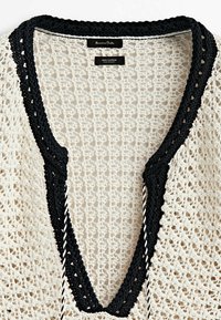 Pull en crochet en coton avec un décolleté en V profond, présentant une bordure noire autour de l'encolure et un tissage texturé ample de couleur crème.