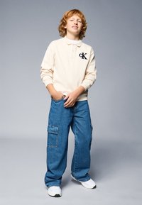 Polo bege com logo, calças cargo azuis de corte relaxado com bolsos laterais, sapatilhas brancas, cabelo cacheado castanho claro, fundo neutro.