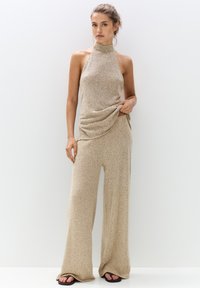 Beige halter-neck jumpsuit in a textured knit fabric, featuring loose wide-leg trousers and draped detailing at the waist.