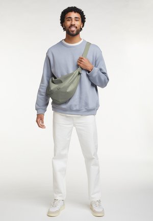 Man in een lichtblauwe trui, witte broek, witte sneakers en een groene crossbodytas, staand en glimlachend tegen een witte achtergrond.