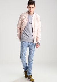 Lys rosa bomberjakke over en grå sweatshirt med sort trykk, svart med lyseblå jeans som har doodle-mønster og grønne sneakers.