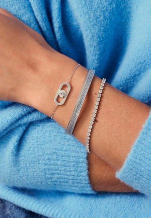 Tre sølvarmbånd på et håndled: et med en central sten, et struktureret, og et med en række små klare sten. Baggrund i blå sweater.