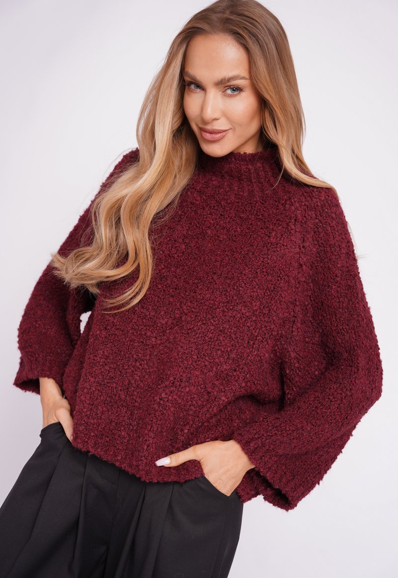 Burgundy textured sweater with a high neckline and loose fit. Features wide sleeves and a cropped hem, paired with black trousers.