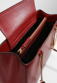 Punainen nahkainen olkalaukku, jossa on teksturoitu pinta, avoin yläosa ja kullanväriset metalliosat. Mukana musta sisusta ja vetoketjullinen tasku. Ketjupidikkeet näkyvissä.