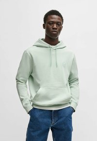 Mint green hoodie in soft fabric with a kangaroo pocket, drawstring hood, and small logo patch on the chest. Paired with blue denim pants.