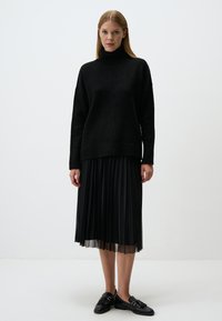 Schwarzer Rollkragenpullover mit lockerer Passform, kombiniert mit einem schwarzen, gefalteten Rock und schwarzen Loafers, vor einem schlichten weißen Hintergrund.