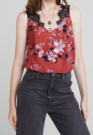 Frau trägt ein rotes ärmelloses Oberteil mit Blumenmuster und schwarzer Spitzenkante sowie mehrere goldene Halsketten, kombiniert mit hoch taillierten schwarzen Jeans aus Denim.