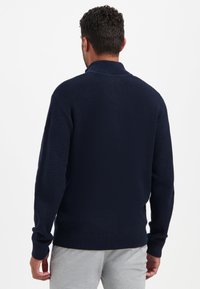 Man in een gestructureerde donkerblauwe gebreide trui en lichtgrijze broek, staande met de rug naar de camera tegen een effen witte achtergrond.