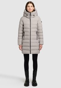 Lys grå dunjakke med høj krave og hætte. Har en frontlynlås, sorte lynlåsdetaljer og sidelommer. Stylet med sorte leggings og støvler.
