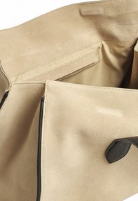 Interno di una borsa in suede beige con una texture liscia, dotata di una tasca laterale e un'etichetta con il marchio. I bordi neri accentuano il design.
