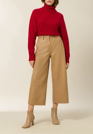 Kobieta ubrana w czerwony sweter z golfem, beżowe szerokie spodnie z wysokim stanem z skóry oraz beżowe botki, stojąca na tle jednolitego tła.