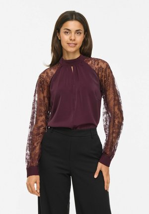 Une femme porte un chemisier couleur prune aux manches en dentelle florale transparente et un pantalon noir sur un fond blanc uni.