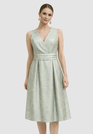 Donna che indossa un abito senza maniche, verde chiaro, lungo fino al ginocchio con motivo floreale e una cintura a tripla fascia, in piedi davanti a uno sfondo uniforme.