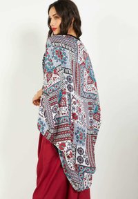 Mujer con cabello ondulado oscuro que lleva una chal multicolor con un patrón floral sobre pantalones rojos, mirando por encima del hombro contra un fondo sencillo.