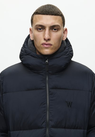 Young man with short hair wears a zipped black puffer jacket with a high collar and a black "W" logo on the chest against a white background.