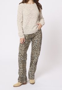 Femme portant un pull en tricot beige, un pantalon à imprimé léopard et des bottines en daim beige, debout avec une main dans la poche.