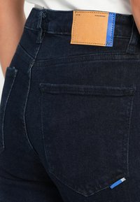 Jeans de denim oscuro con un acabado texturizado, que cuentan con un parche de cuero marrón claro con acentos azules, bolsillos traseros visibles y detalles de costura estándar.