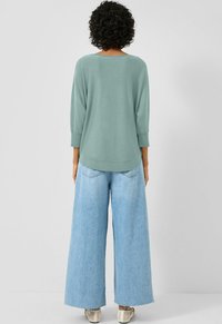 Osoba z kręconymi włosami w jasnoszarym, długim topie z długimi rękawami, w szerokich, jasnoniebieskich dżinsach i białych płaskich butach, stojąca na jednolitym tle.