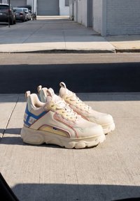 Klobige beige Sneakers mit dicken Sohlen und blauen und gelben Akzenten, die bei hellem Sonnenlicht auf einem Betonweg stehen.