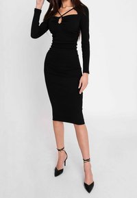 Donna con un vestito midi nero aderente con maniche lunghe e scollatura incrociata, abbinato a tacchi alti a punta neri con lacci alla caviglia.