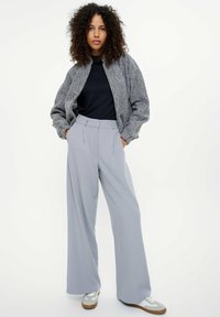 Kobieta z kręconymi włosami ubrana w szaro-zieloną kurtkę na zamek, czarną bluzkę, szerokie jasnoszare spodnie oraz białe trampki, stoi z rękami w kieszeniach.