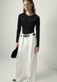 Mujer vistiendo una blusa negra de manga larga con cuello ancho, pantalones blancos de tiro alto y pierna ancha, sosteniendo un pequeño bolso negro, de pie en un entorno simple.