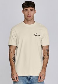 Camiseta de algodón color crema con cuello redondo y mangas cortas, con un logo en cursiva negra en el lado izquierdo del pecho.