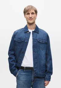 Τζάκετ από τζιν σε σκούρο μπλε χρώμα με κουμπιά μπροστά, δύο τσέπες στο στήθος και κλασικό γιακά. Φοράει πάνω από ένα λευκό T-shirt.