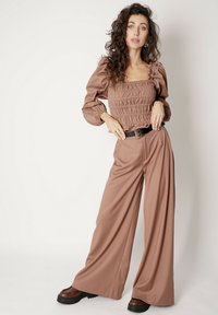 Jasnobrązowa, marszczona bluzka z długim rękawem zestawiona z szerokimi spodniami w dopasowanym kolorze. Szczegóły obejmują czarny pasek i masywne brązowe buty na platformie.