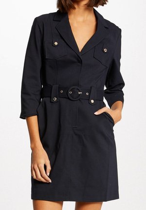 Femme portant une robe noire ceinturée avec col à revers, manches trois-quarts, poches poitrine à rabat avec boutons, et poches latérales.