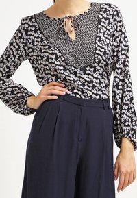 Blusa de manga larga en negro con patrón floral blanco, con escote en V, cierre con lazo y panel insertado texturizado. Combinada con pantalones azul marino.