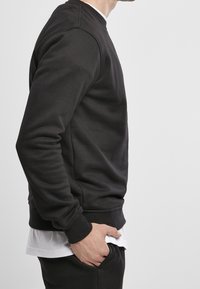Homme portant un sweat-shirt noir sur une chemise blanche, main dans la poche, vu de profil sur un fond clair uni.
